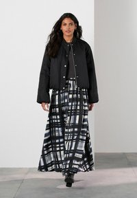 Vrouw loopt in een zwart bomberjack over een zwart top en een lange zwart-witte geruite rok met zwarte haklaarzen.