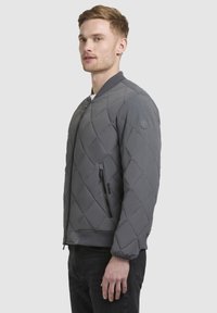 Jachetă bomber matlasată gri, cu un buzunar cu fermoar, mansete și tiv ribbate, având un logo pe mânecă. Textură netedă și captusită.