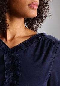 Marinblus med en mjuk, slät textur, dekorativ volang vid halsringningen och knappslån. Designen har en grund V-hals och subtila veck.