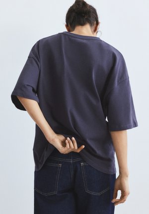 Osoba s tamnom kosom u širokoj tamnoplavoj majici i trapericama, okrenuta leđima, s jednom rukom koja povlači rub majice na leđima.