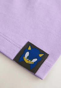 Lys lilla bomuldsstof har en sort lap med et blåt tegneseriefigur ansigt, inklusive grønne øjne og gule accenter.