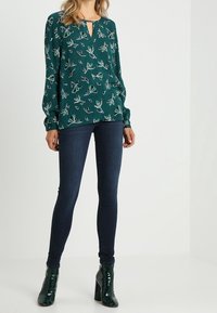 Blusa floral verde con mangas largas, escote en forma de llave, combinada con vaqueros ajustados oscuros y botas de tacón alto negras.