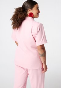 Lichtroze twee-delige outfit met een shirt met korte mouwen en bijpassende broek. Zachte stof met een comfortabele pasvorm; zichtbare naaddetails op de rug.