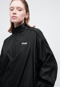 Kvinde iført løs sort zip-up jakke med "ROSIE" og Puma-logo på brystet, poseret mod en ensfarvet lys baggrund.