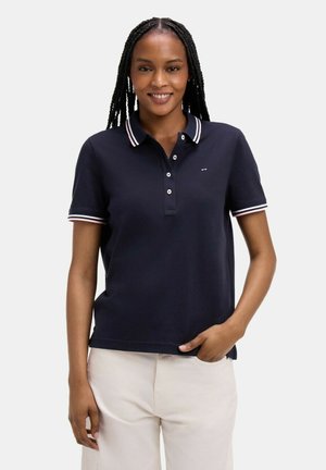 Femme aux longues tresses portant un polo bleu marine avec des finitions blanches et rouges et un pantalon beige clair, souriante avec la main dans la poche.
