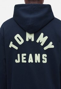 Човек, носещ тъмносин суичър с големи бели букви, които гласят "TOMMY JEANS" на гърба във формата на арка.