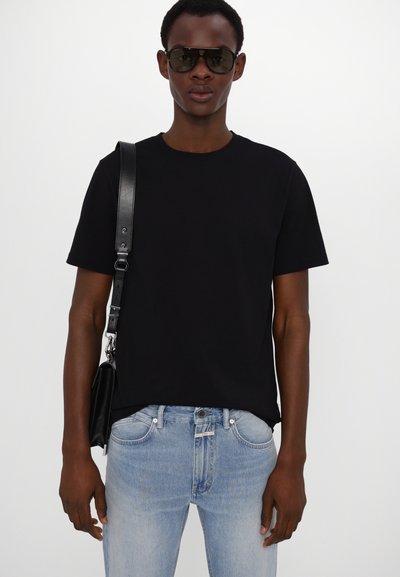 T-shirt noir à col ras du cou en coton, associé à un jean en denim bleu clair. Le mannequin tient un sac noir avec une fine sangle en cuir et porte des lunettes de soleil.