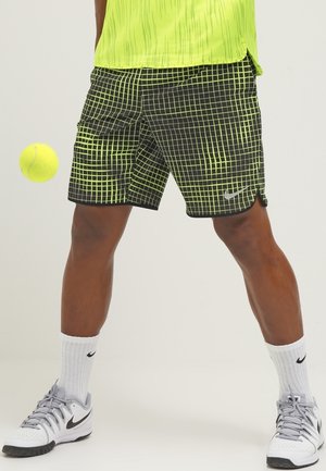 Person trägt schwarz-neongrüne Shorts mit Karomuster, gelbes Oberteil, weiße Nike-Socken und Schuhe, mit einem Tennisball in der Nähe des linken Beins.