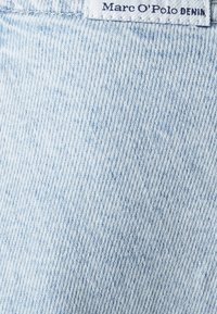 Lys blå denim-stoff med synlig struktur og lett falming. Har en hvit etikett på toppen som står "Marc O'Polo DENIM."