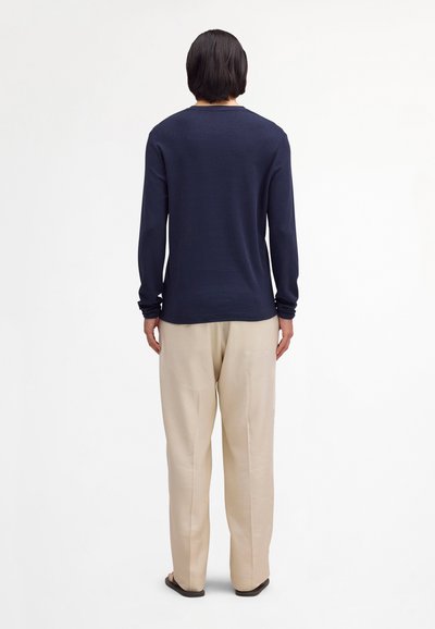 Osoba s krátkymi čiernymi vlasmi, oblečená v tmavomodrej dlhoruká košeli, béžových nohaviciach a čiernych topánkach, zobrazená zozadu na bielom pozadí.