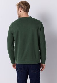 Dunkelgrüner Sweatshirt mit Rundhalsausschnitt, langen Ärmeln, gerippten Bündchen und geradem Saum. Kombiniert mit dunkelblauen Jeans.