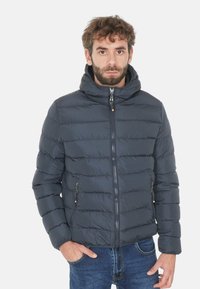 Giacca puffer blu scuro con cappuccio, caratterizzata da un design trapuntato, chiusura frontale con zip e due tasche laterali con zip. Realizzata in materiale leggero.