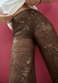 Sheer brown pants with floral lace embroidery and sequin accents, featuring a fitted waist and flared legs against a pink background.