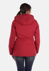 Giacca isolante rossa con cappuccio, presenta una forma sagomata, una texture liscia e dettagli di cucitura visibili, indossata con jeans scuri.