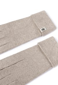 Beige gestrickte Handschuhe mit umgeschlagenen Bündchen und einem kleinen schwarz-weißen Logosticker an einem Bündchen, nebeneinander auf einem weißen Hintergrund abgebildet.