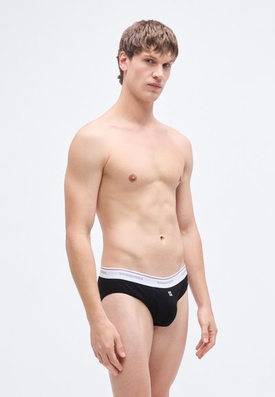 Slip noir avec ceinture élastique blanche portant la marque "DSQUARED2", coupe ajustée et texture lisse, porté par un homme debout.