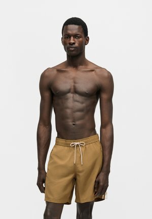 Tummaihoinen, lihaksikas mies, jolla on beige väriset uimashortsit, jotka sidotaan narulla, seisoo yksinkertaisen valkoisen taustan edessä.