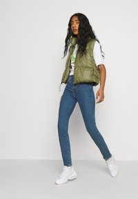 Chaleco acolchado verde oliva sobre una camiseta gráfica blanca, combinado con jeans ajustados azules y zapatillas blancas, exhibido sobre un fondo neutro.