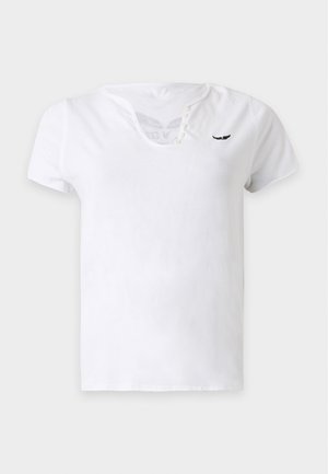 Femme portant un t-shirt blanc cintré à col en V, jean bleu clair, bottes en daim marron, tenant un sac à main en cuir marron sur un fond clair uni.