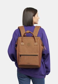 Zaino in tela marrone con manico superiore e spallacci regolabili, dotato di una tasca frontale con zip e di un logo circolare sul retro.