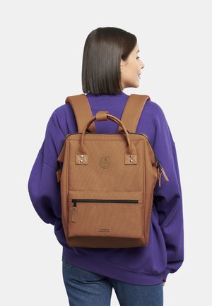 Zaino in tela marrone con manico superiore e spallacci regolabili, dotato di una tasca frontale con zip e di un logo circolare sul retro.