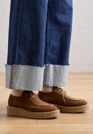 Chaussures à lacets en daim marron portées avec des chaussettes claires et un jean bleu large avec revers, debout sur un sol en bois.