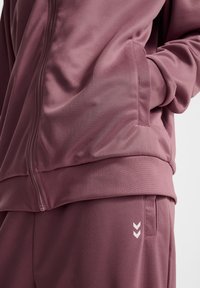Mauve atletisk zip-up jakke med sidelommer og matchende bukser, med en glat tekstur og hvide chevron detaljer på benet.