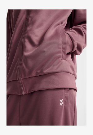 Mauve Sportjacke mit Reißverschluss, Seitentaschen und passenden Hosen, die eine glatte Textur und weiße Chevron-Akzente an den Beinen aufweisen.