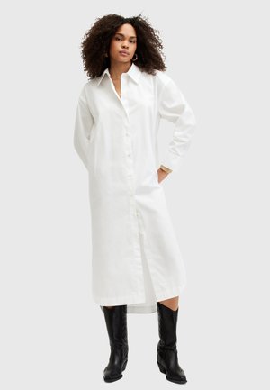 Vestido blanco de manga larga con botones en la parte delantera, cuello con solapa, bolsillos laterales y un corte relajado, combinado con botas negras hasta la rodilla.