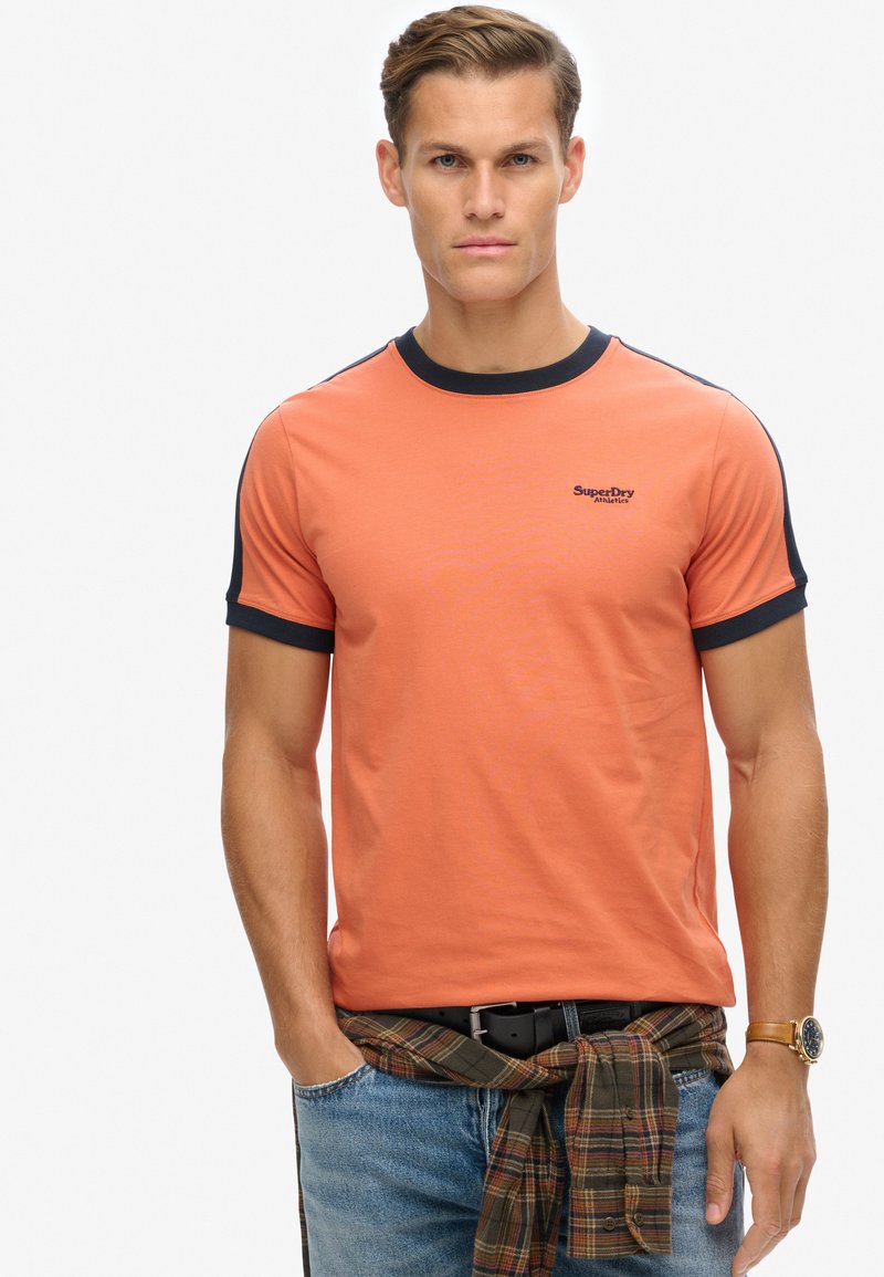 Orange bomulds t-shirt med marineblå ribbet krave og ærmer, der har et lille SuperDry-logo. Modellen har blå jeans på og en ternet skjorte bundet om livet.