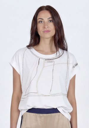 Hosszú barna hajú nő fehér, mintás rövid ujjú ingben és bézs nadrágban, egyszerű fehér háttér előtt áll.