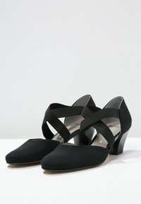 Scarpe con tacco nere con tomaia in tessuto morbido. Presentano cinturini elastici incrociati e una punta arrotondata. Tacco basso a blocco.