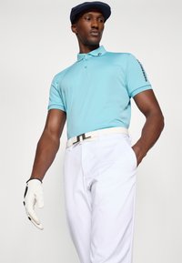 Lyseblå polo skjorte med krage og emblemet på ermet, kombinert med hvite bukser. Et hvitt belte og hvit golfhanske fullfører antrekket.