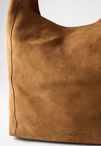 Brown suede handbag with a soft texture, minimalistic design, and visible stitching details. Features a stamped logo on the side.
