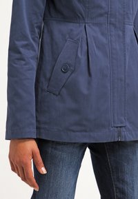 Marinblå lättviktsjacka med plisserad design, knappad sidoficka och dragkedjestängning framtill. Slät, strukturerad tyg.