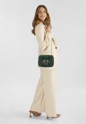 Vrouw in een crème broekpak en hakken, die een donkergroene gewatteerde handtas met kettinghengsel en metalen logo vasthoudt, glimlacht en over haar schouder kijkt.