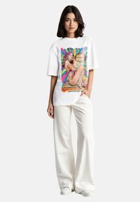 Witte oversized t-shirt met een kleurrafische weergave van een zittende vrouw met de tekst "Good Vibes." Gecombineerd met wijde crèmekleurige broek en witte schoenen.