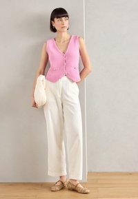 Chaleco de punto rosa con escote en V y cierre de botones, combinado con pantalones de pierna ancha blancos con acanalado y zapatos planos de colores con patrón. Sosteniendo un bolso crema.