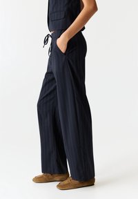 Pantalon large bleu marine en tissu léger, présentant un motif discret à fines rayures, des poches avant et une taille réglable avec cordon de serrage.