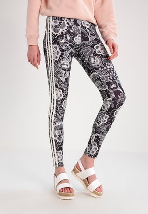 Zwarte leggings met een bloemenkantpatroon, voorzien van witte zijstrepen. Gecombineerd met witte platform sandalen en een lichtroze trui.