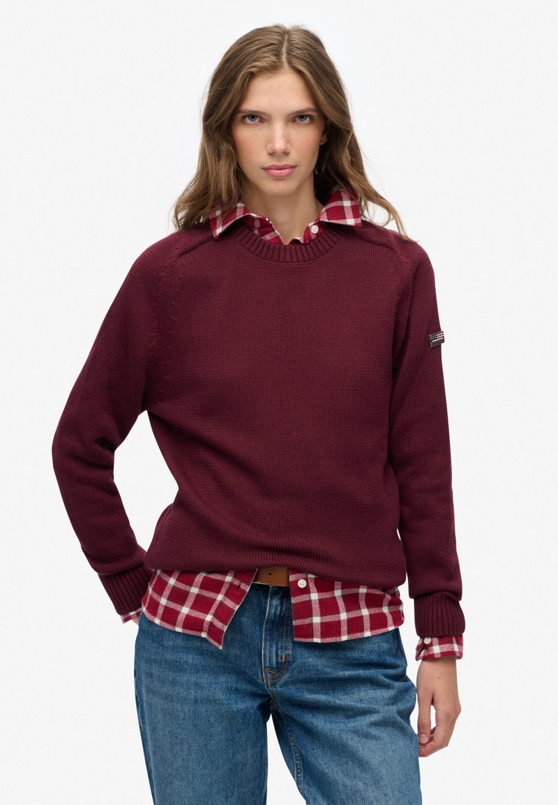 Burgundy knit sweater with a round neckline and ribbed cuffs, layered over a red and white plaid shirt, paired with blue jeans.