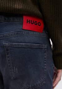 Mørkeblå denimjeans med en strukturert finish, med en fremtredende rød lærmerke trykt med "HUGO" i svart. To baklommer.