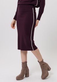 Knie-lange nauwsluitende rok in dieppaars gebreid met een contrasterende witte zijstreep. Gecombineerd met taupekleurige suède enkellaarzen.