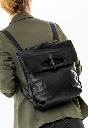Persoon draagt een olijfgroene jas en een grote zwarte leren rugzak met een klep en gespsluiting.