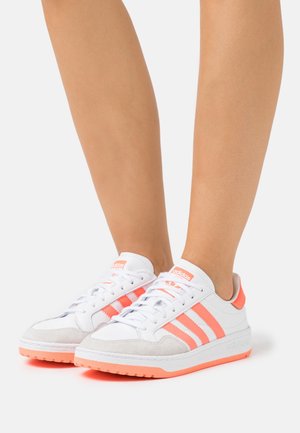 Baskets blanches avec accents roses, bout en daim, et trois bandes roses sur les côtés, dotées d'une semelle en caoutchouc texturée et de lacets blancs.