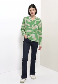 Blusa verde a maniche lunghe con motivi astratti beige, abbinata a jeans a zampa blu scuro e sneaker bianche. Tessuto liscio, vestibilità rilassata.