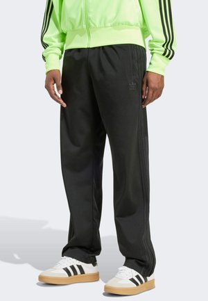 Person trägt schwarze Adidas-Trainingshose, weiße Sneaker mit schwarzen Streifen und eine neongrüne Jacke mit schwarzen Streifen an den Ärmeln
