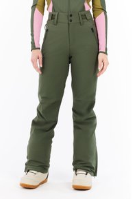 Woman wearing green snow pants with zippered pockets and beige snow boots, standing against white background.