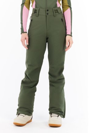 Frau in grünen Schneehosen mit Reißverschlusstaschen und beigen Schneestiefeln, die vor einem weißen Hintergrund steht.