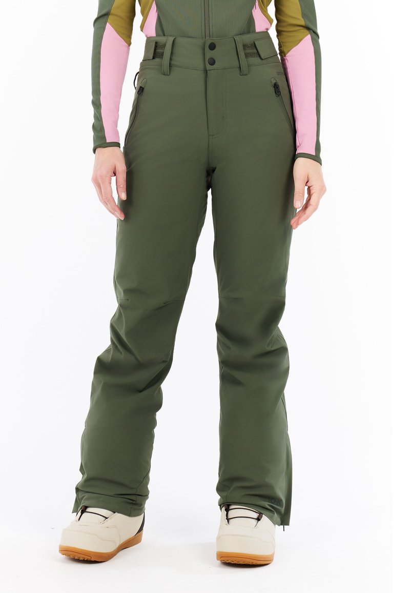 Woman wearing green snow pants with zippered pockets and beige snow boots, standing against white background.